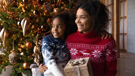 Close up dreamy African American woman with adorable daughter looking to aside, holding gift box,...