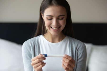 Awaited happiness. Excited millennial woman sitting on bed at home in the morning studying positive result of pregnancy test with happy smile getting planning expected gestation successful ivf attemptの写真素材