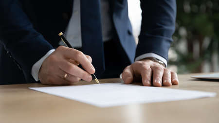 Close up young businessman standing near table with pen in hands, ready signing profitable offer agreement after checking contract terms of conditions, executive manager involved in legal paperwork.の写真素材