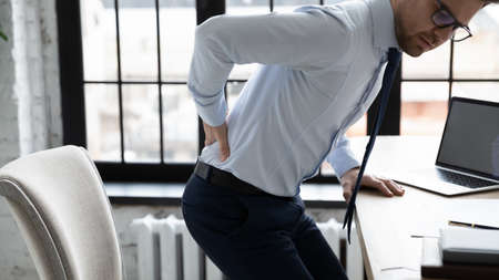 Unhealthy young male office worker getting up from chair, having painful feelings in lower back, weak sick unhappy millennial entrepreneur businessman suffering from backpain touching muscles.の写真素材