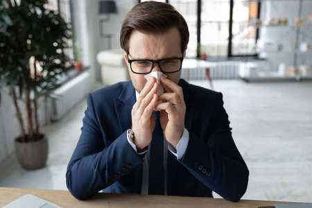 Unhealthy millennial businessman suffering from runny nose using paper handkerchief in office,...