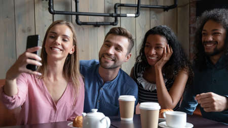 Hello, here we are. Joyful company of young diverse mates millennial men and women posing for group selfie sitting close together at table in cafe, making video call by cellphone to absent friendの写真素材