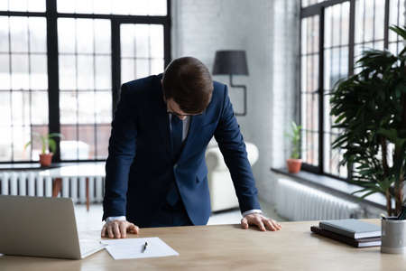 Frustrated young businessman in formal wear standing at office table with head down, feeling depressed after professional failure, company bankruptcy, money loss or unprofitable deal concluding.の写真素材