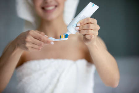 Easy way to healthy smile. Close up of smiling young female standing at bathroom in towel around body applying paste on toothbrush preparing to clean teeth following oral hygiene looking after herselfの写真素材