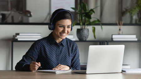 Smiling Indian businesswoman intern wearing headphones watching webinar, writing notes, listening to...