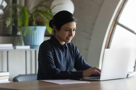 Confident Indian businesswoman entrepreneur working on laptop in modern office, looking at screen, typing, focused smiling female employee worker writing financial report, online projectの写真素材