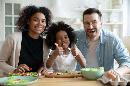 Head shot portrait happy multiracial family with kid cooking in kitchen, smiling African American mother and Caucasian father with adorable little daughter showing thumbs up looking at cameraの写真素材