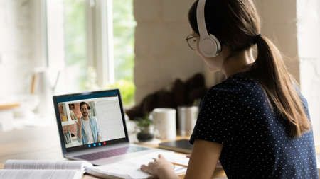 Rear back view happy young caucasian girl in headphones holding video call consultation with skilled...