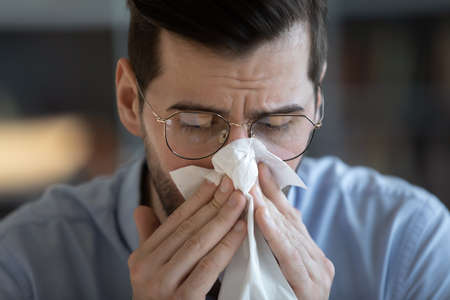 Close up young 35s man in glasses holding tissue and sneezing, blowing runny nose in paper...