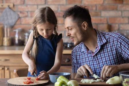 Am I doing it right, daddy. Caring single father teaching attentive little daughter cook meals at kitchen, showing how to cut vegetables correct and hold knife safe, giving advice, explaining recipeの写真素材