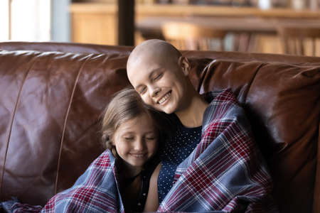 Enjoying every moment together. Tender loving mother or elder sister struggling with oncology disease sitting on sofa with cute small daughter child embracing and covering shoulders by warm cozy plaidの写真素材