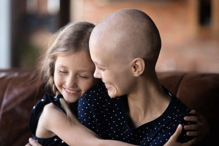 Sense of life. Tender loving young single mother cancer patient and little girl daughter of school age cuddling on sofa at living room holding each other in arms supporting caring giving strength hopeの写真素材