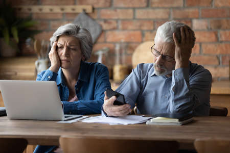 Unhappy mature couple checking financial documents, calculating domestic bills, having problem with money, upset senior man wearing glasses and woman feeling depressed about bankruptcy or debtの写真素材