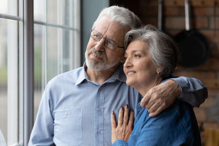 Happy dreamy mature couple hugging, thinking about good future together, standing in kitchen,...