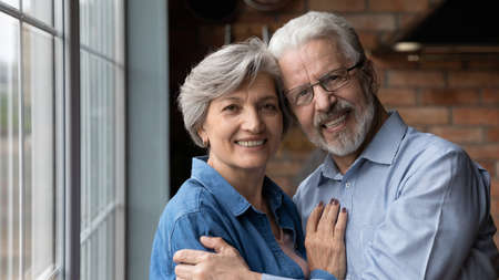 Head shot portrait smiling mature couple hugging, looking at camera, standing near window in kitchen...