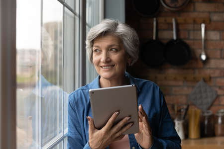 Close up smiling dreamy mature woman holding tablet, standing near window at home, happy grey haired female distracted from chatting online in social network, waiting for video call from relativesの写真素材
