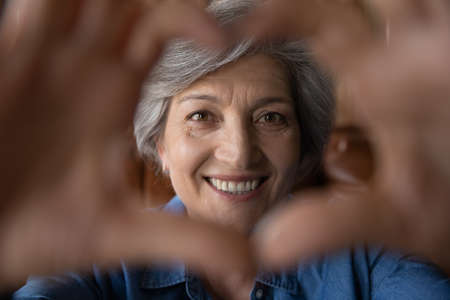 Close up head shot portrait smiling mature grey haired woman showing heart gesture, looking through...