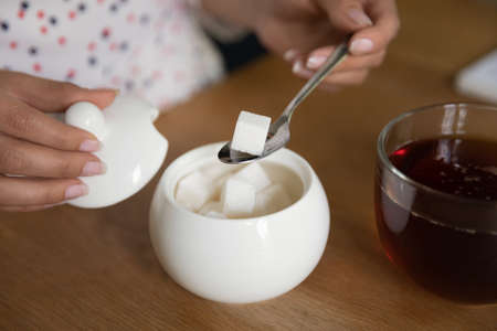 Crop close up of woman put cube of white sugar drinking warm hot coffee at home. Female tenant enjoy...