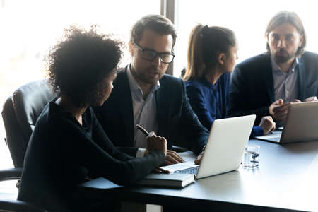 Serious multiracial businesspeople gather in office work in groups using laptops discussing ideas. Concentrated diverse colleagues coworkers look at computer screen engaged in teamwork at briefing.の写真素材