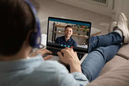 Rear shoulder view relaxed young man in earphones holding video call online web meeting with...