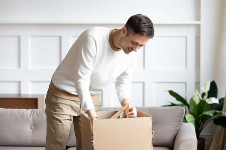 Man standing in living room on day of move to rented home, unbox carton box with belongings. Relocate to new own apartment collect personal things. Delivered parcel to satisfied client, tenant conceptの写真素材