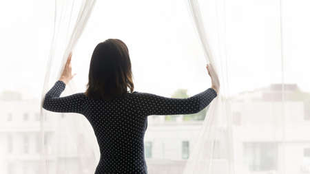 Rear view beautiful Asian young woman opening curtains in morning, attractive female dreaming, starting new day, planning good future, enjoying sunlight, standing near window at homeの写真素材