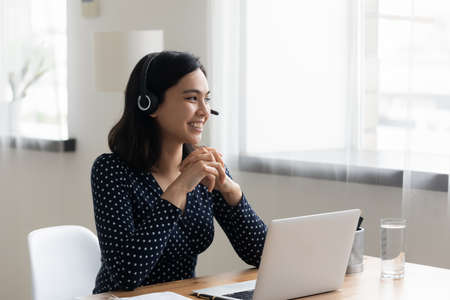 Smiling Asian businesswoman wearing headset using laptop, looking to aside, dreaming about new job opportunities, happy woman student watching webinar, listening to lecture, studying, home officeの写真素材