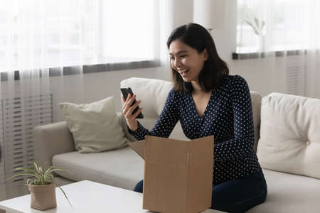 Overjoyed Asian woman unpacking parcel, using phone, excited female vlogger shooting unboxing video,...