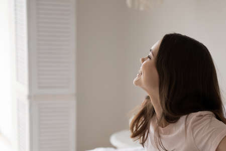 Hello, world. Happy smiling millennial female waking up at home or in hotel suite in perfect mood, sitting on bed looking at window glad to meet morning sun, planning thinking of spending new good dayの写真素材