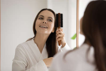 Excellent result. Happy excited young lady brushing hair after shower looking at big mirror satisfied with perfect condition of long shiny strands after regular using of revitalizing shampoo balm maskの写真素材