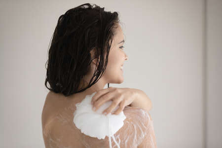 Enjoying pleasant moment. Rear view of happy young lady relaxing in shower, having pleasure standing under warm running water, rubbing herself with soft bath puff with perfumed foamy gel or cream soapの写真素材