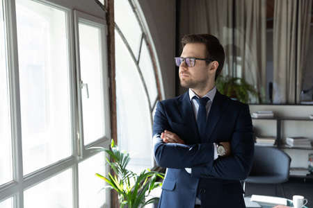 Pensive young company owner head of department standing by panoramic office window holding arms crossed on chest proud of personal achievement planning business expansion visualizing wellbeing successの写真素材