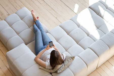 Top view woman using smartphone, relaxing on couch in living room, young female holding phone with...