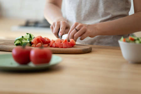 Close up woman cooking salad, cutting organic fresh vegetables, cucumbers and tomatoes with knife on board, standing at kitchen table, young female preparing lunch, vegetarian mealの写真素材