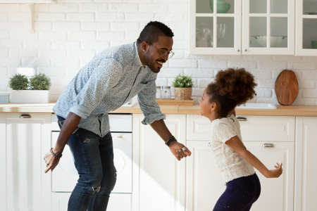 Funny cooking with dad. Happy laughing millennial black father dancing disco at modern designed kitchen with overjoyed little kid daughter jumping listening to music enjoying active leisure having funの写真素材