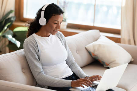 Millennial biracial woman in headphones sit relax on couch at home work online type text on laptop....