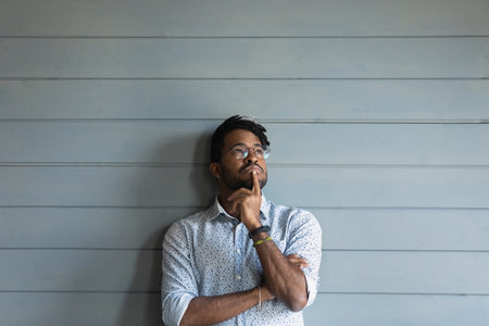 Thoughtful young african american man looking up at copy space for advertisement text, posing near wall. Pensive 30s mixed race guy in eyeglasses making decision, isolated on grey background.の写真素材