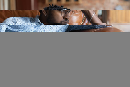 Frustrated young african ethnicity man in glasses sitting on sofa, thinking or life problems, feeling stressed or disappointed alone at home. Thoughtful millennial mixed race guy looking in distance.の写真素材