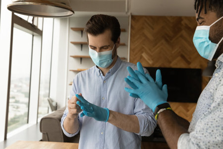 Smiling multiracial male colleagues put rubber gloves and medical face masks protect from...