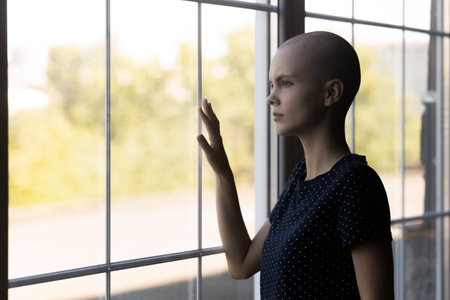 Close up unhappy sick hairless woman lost in thoughts, standing near window, touching glass, looking to aside, thinking about health problem, dreaming about recovery and remission, futureの写真素材