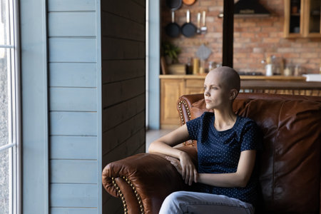 Thoughtful hairless sick woman sitting in armchair alone, looking in distance, lost in thoughts, pensive female oncology cancer patient dreaming about recovery and remission, visualizing futureの写真素材