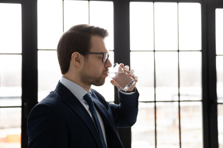 Side view confident businessman wearing glasses drinking fresh pure mineral water, standing near window, employee preventing dehydration, skin and health care, healthy lifestyle habit conceptの写真素材