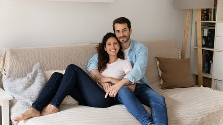 Portrait of young wife and husband hugging, sitting on couch, smiling attractive woman and man cuddling, looking at camera, posing for family photo at home, relaxing on sofa, enjoying lazy weekendの写真素材
