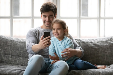Smiling handsome young father embracing adorable small kid daughter, looking at smartphone web camera, positing for selfie photo, recording video or holding distant videocall, sitting on sofa.の写真素材