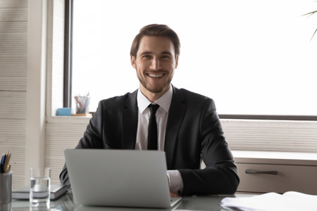 Portrait of smiling young successful Caucasian businessman sit at desk in office work on laptop. Happy millennial male CEO or director look at camera show confidence and leadership at workplace.の写真素材