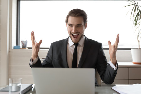 Overjoyed young Caucasian businessman feel euphoric read good amazing news online on laptop. Excited male CEO sit at desk in office triumph get business win on computer. Luck, success concept.の写真素材