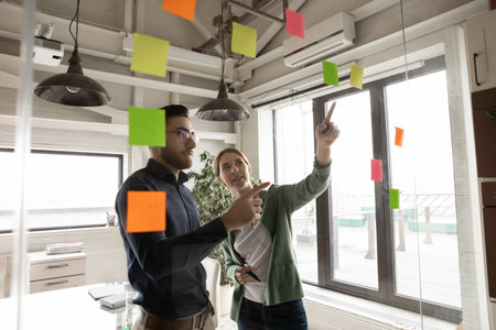 Motivated multiracial colleagues look at glass office wall work with colorful sticker note pads planning together. Multiethnic employees engaged in creative thinking in boardroom. Teamwork concept.の写真素材