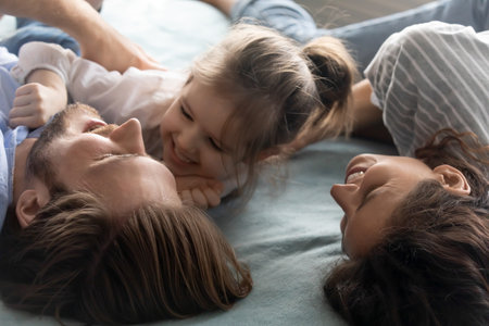 Close up of overjoyed young Caucasian parents lying in bed have fun play with little daughter....