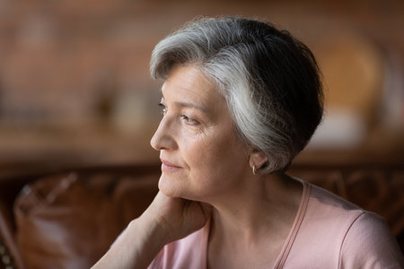 Head shot close up thoughtful happy middle aged mature woman looking in distance, relaxing on sofa, recollecting good memories or enjoying peaceful moment alone indoors, domestic meditation concept.の写真素材