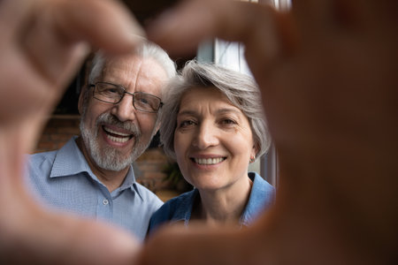 Head shot close up happy bonding older senior family couple making heart sign with fingers, showing...